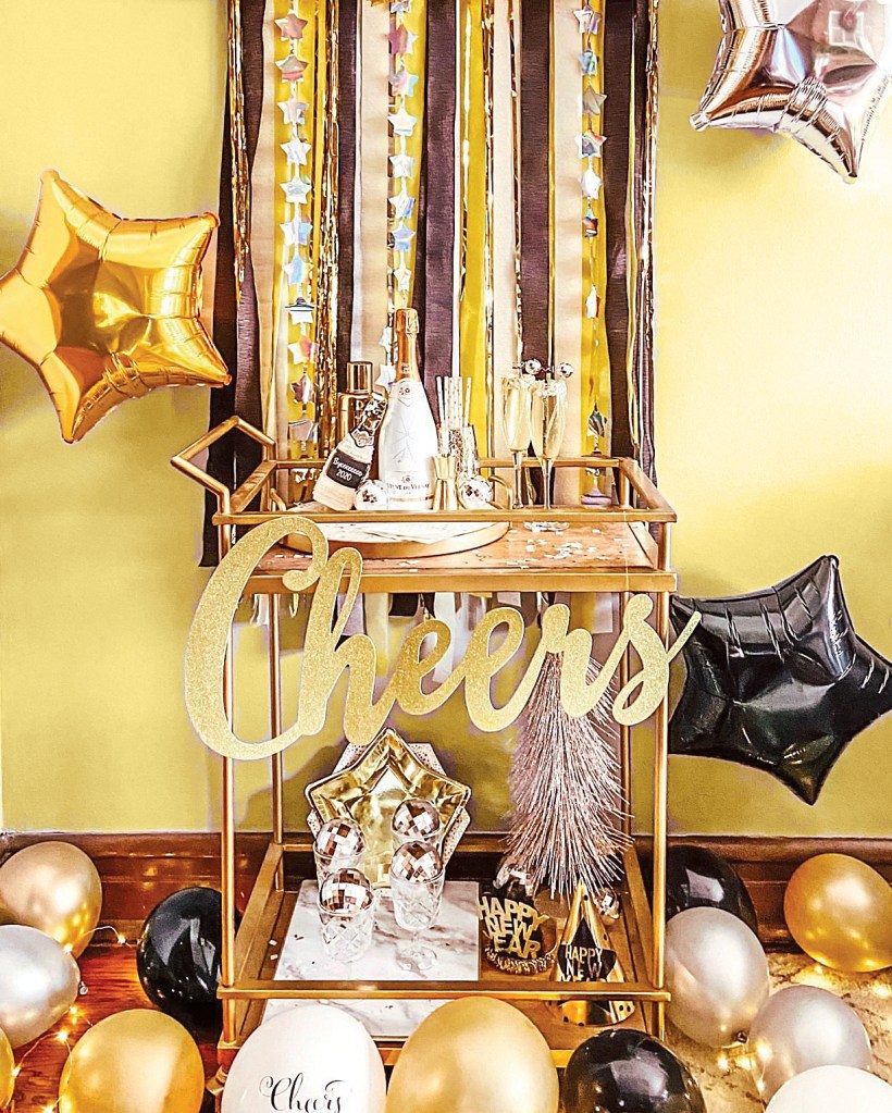Bar cart decor idea featuring a glam gold champagne flute and bottle-filled cart positioned in front of sparkly gold, black and silver party streamers attached to wall