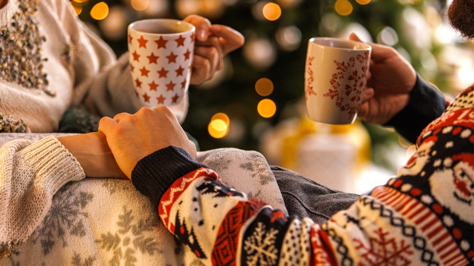 Holiday romance books: Featured image of couple with cocoa cuddling on couch by Christmas tree