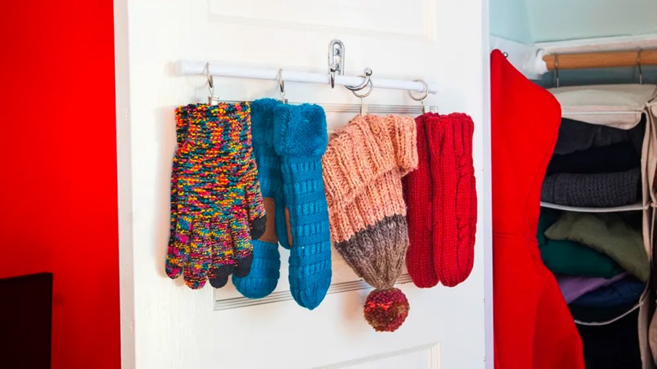 Winter hats and gloves hung from shower hooks on a tension rod attached to the back of a white closet door with a mounted hook