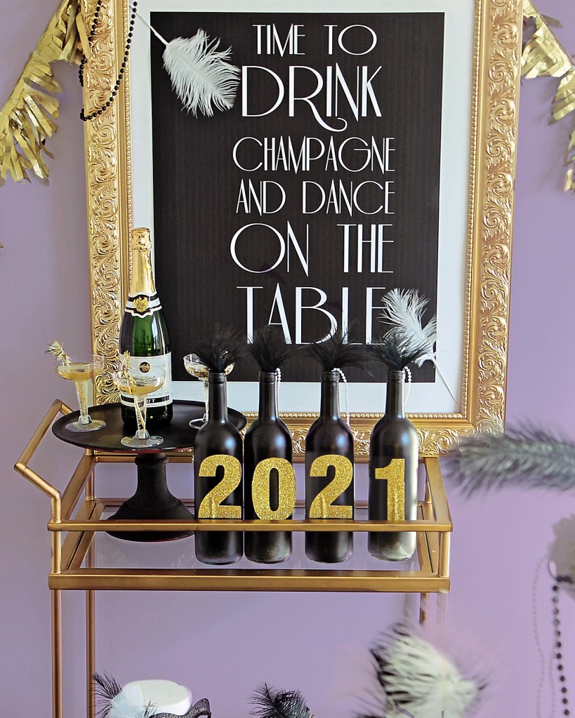 Bar cart decor idea featuring a gold bubbly bar cart decorated with champagne flutes and disguised bottles of bubbly and positioned in front of a festive 1920's themed framed sign 