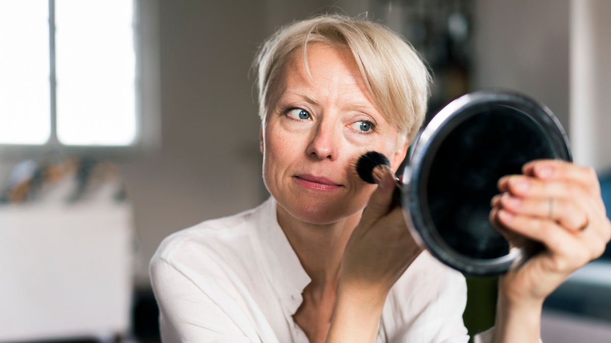 Mature woman applying blush to skin in mirror