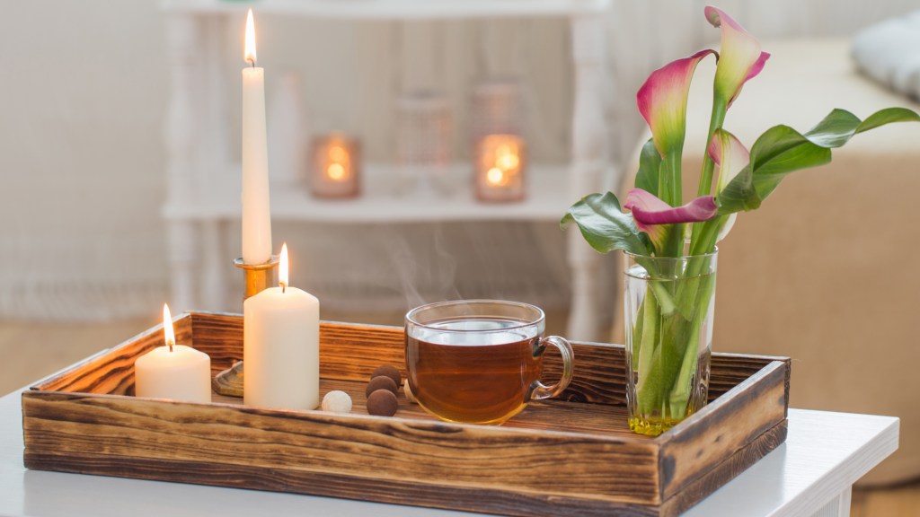 How to decorate a coffee table: White coffee table topped with a wooden tray that's filled with three candles, a mug of tea, chocolate truffles and drinking glass of pink lilies
