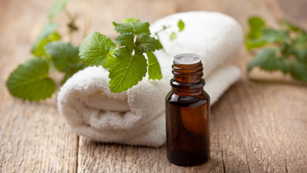 An amber vial of mint essential oil beside a fresh sprig of mint on a while towel, which is used to treat a tension headache