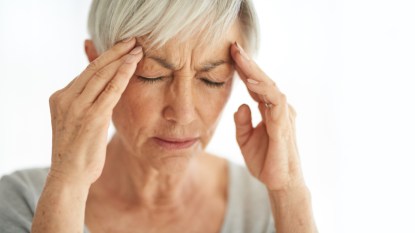 A woman with grey hair and closed eyes, holding her hands to her head with a tension headache or migraine