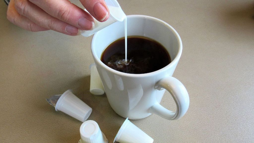 Coffee creamer being poured into a cup of black coffee, from a small, disposable, plastic container.
