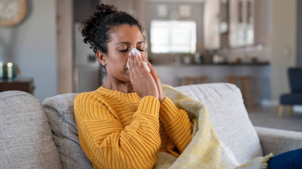 A woman in a yellow sweater sitting on a couch, closing her eyes as she blows her nose into a tissue