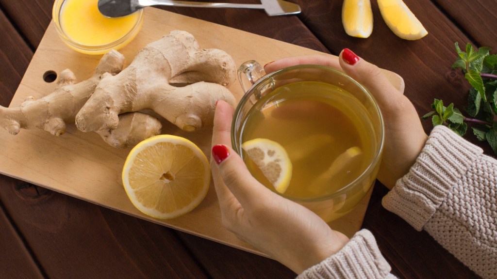 A close up of a woman with red nails holding a cup of ginger tea for bloating next to fresh ginger and lemon slices