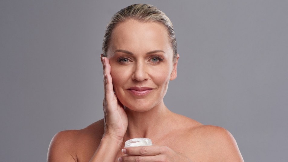Mature woman smiling with hand on face and holding moisturizer for rosacea