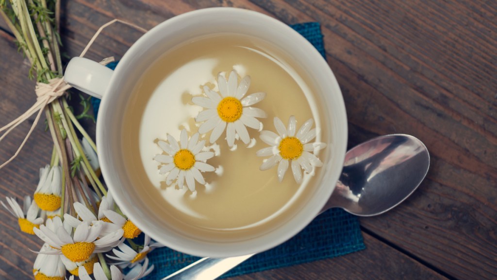 A mug of chamomile tea for bloating beside fresh chamomile flowers and a spoon