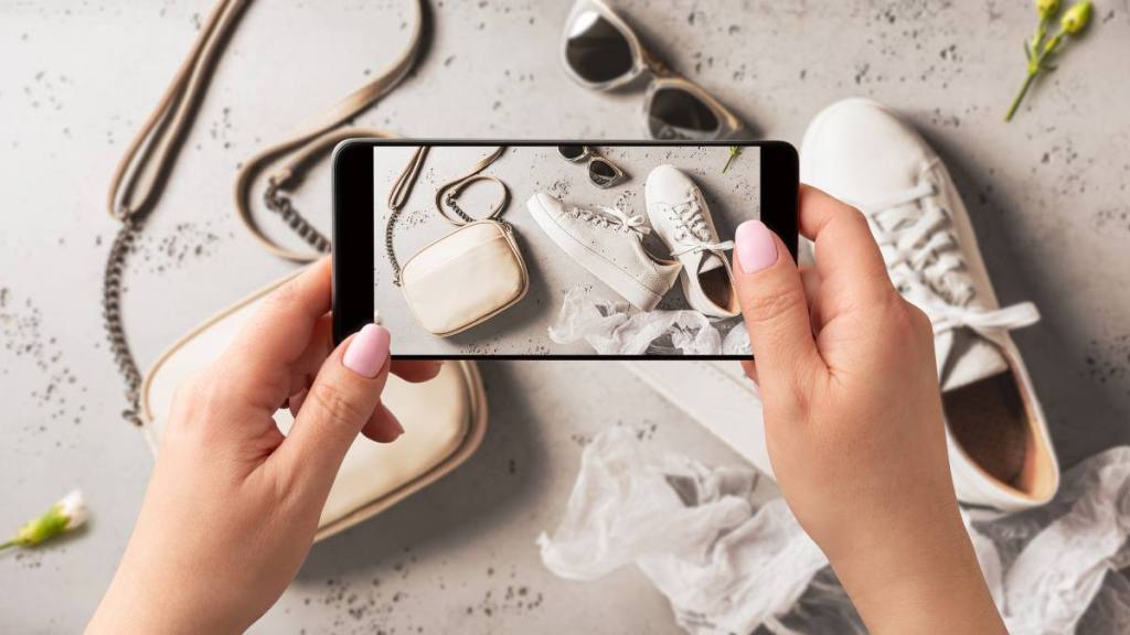 how to sell stuff online: Woman taking photo of white leather sneakers shoes, handbag and sunglasses with smartphone. Blogger, influencer or stylist capturing spring fashion accessories for social media. Grey background.