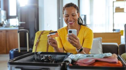 save on spring travel: Mature African American woman packing for a trip and using online payment services