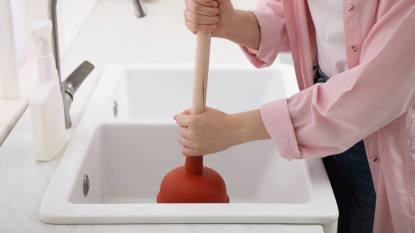 How to unclog kitchen sink: Woman using plunger to unclog sink drain in kitchen, closeup