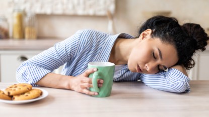 A woman with dark hair asleep at a table after eating while holding a cup of coffee