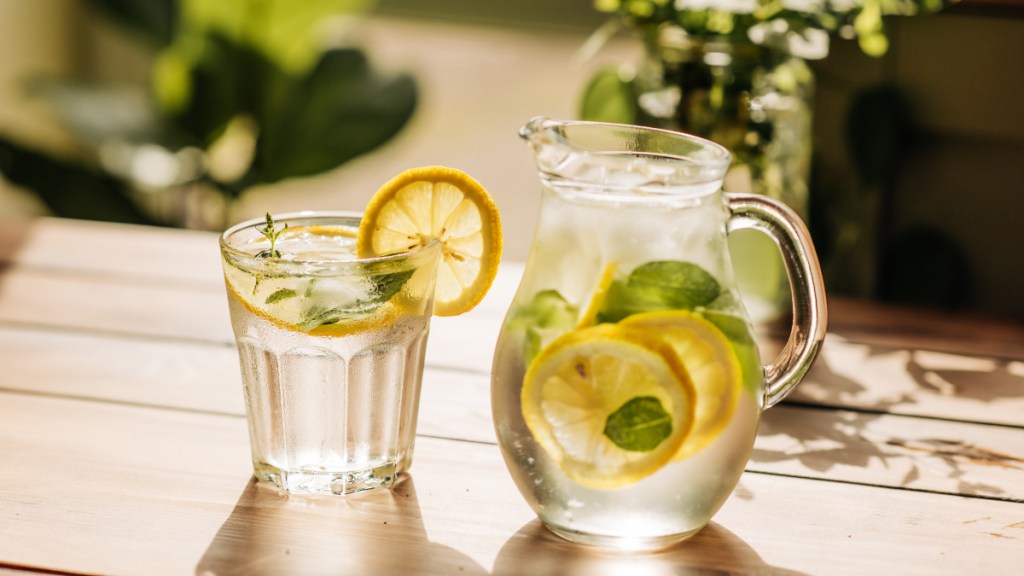 A pitcher of water with lemon slices and herbs beside a glass of water on a table