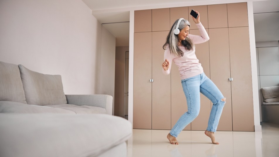 cheerful lady in wireless headphones holding mobile phone and moving to the rhythm of music while having fun in living room
