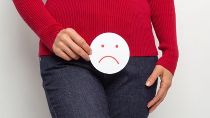A close-up of a woman in a red top and jeans holding a frowning face in front of her to signal a vaginal ulcer