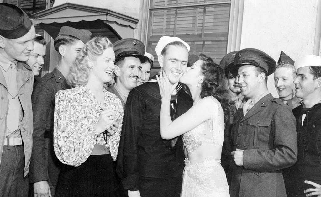 Yvonne De Carlo plants a resounding kiss on S1/C Harold Farnlund, Mountain Home, Idaho, as other servicemen wait their turn. Yvonne had to kiss the first 50 servicemen she met on Hollywood Boulevard and actress Martha O'Driscoll (Left) keeps tab of the payoff