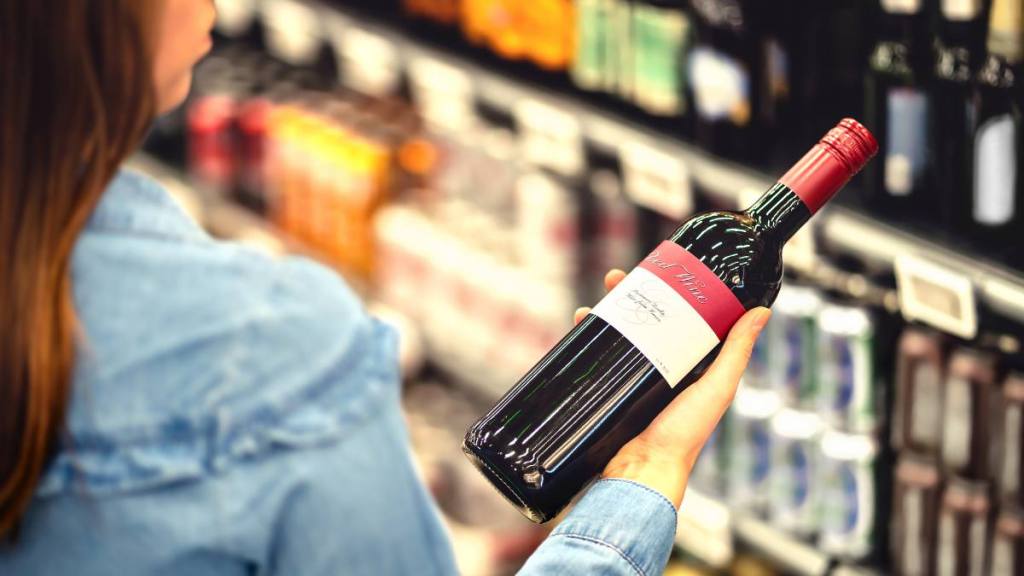 Gifts for Mother's Day: Woman reading the label of red wine bottle in liquor store or alcohol section of supermarket. Shelf full of alcoholic beverages. Female customer holding and choosing a bottle of merlot or sangiovese.