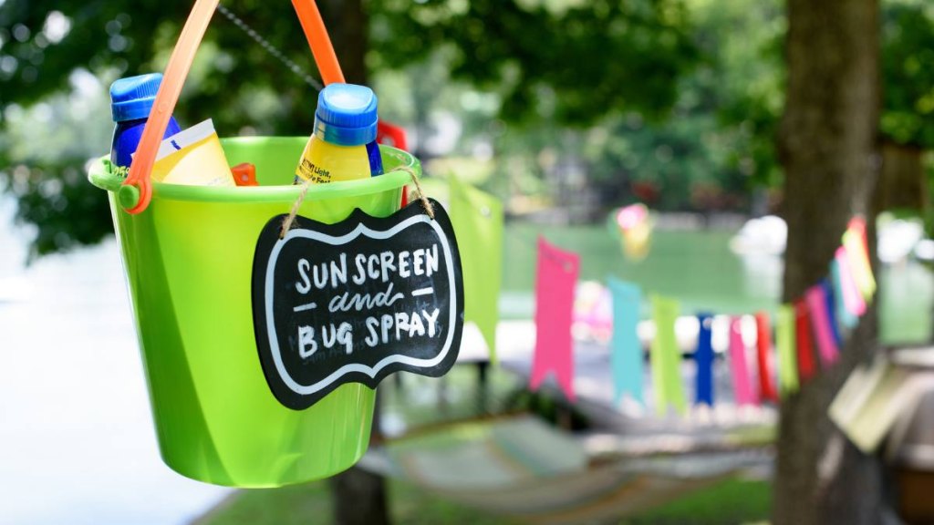 Sunscreen and bug spray in a green bucket with a handwritten chalk sign on the front and party decorations nearby (babs bbq tips)