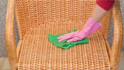 how to clean wicker furniture: Woman's hand in pink rubber protective glove wiping bamboo straw armchair from dust with dry rag. Early spring cleaning or regular clean up. Maid cleans house.