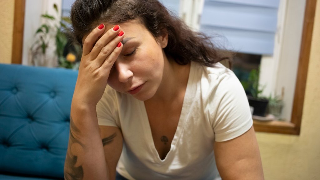 A woman sits on the couch with a hand on her head in pain
