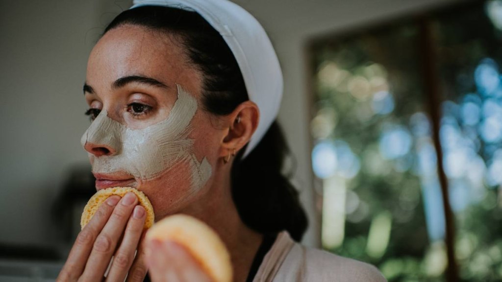 A woman removes a clay face mask from her face using two, small, reusable yellow sponges.