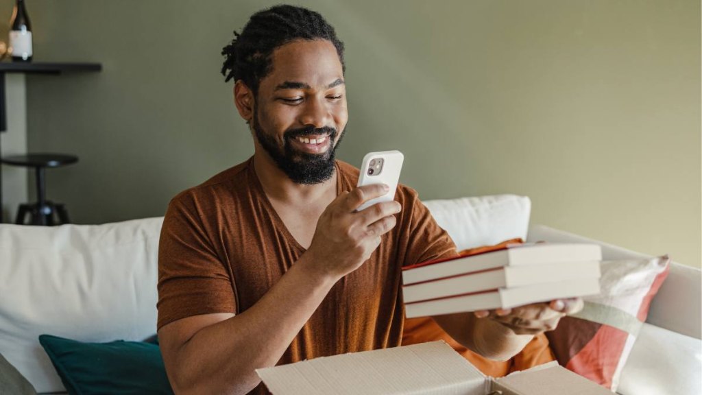 Young African American man is at home. He is unpacking the delivered package and taking a picture of the ordered item.