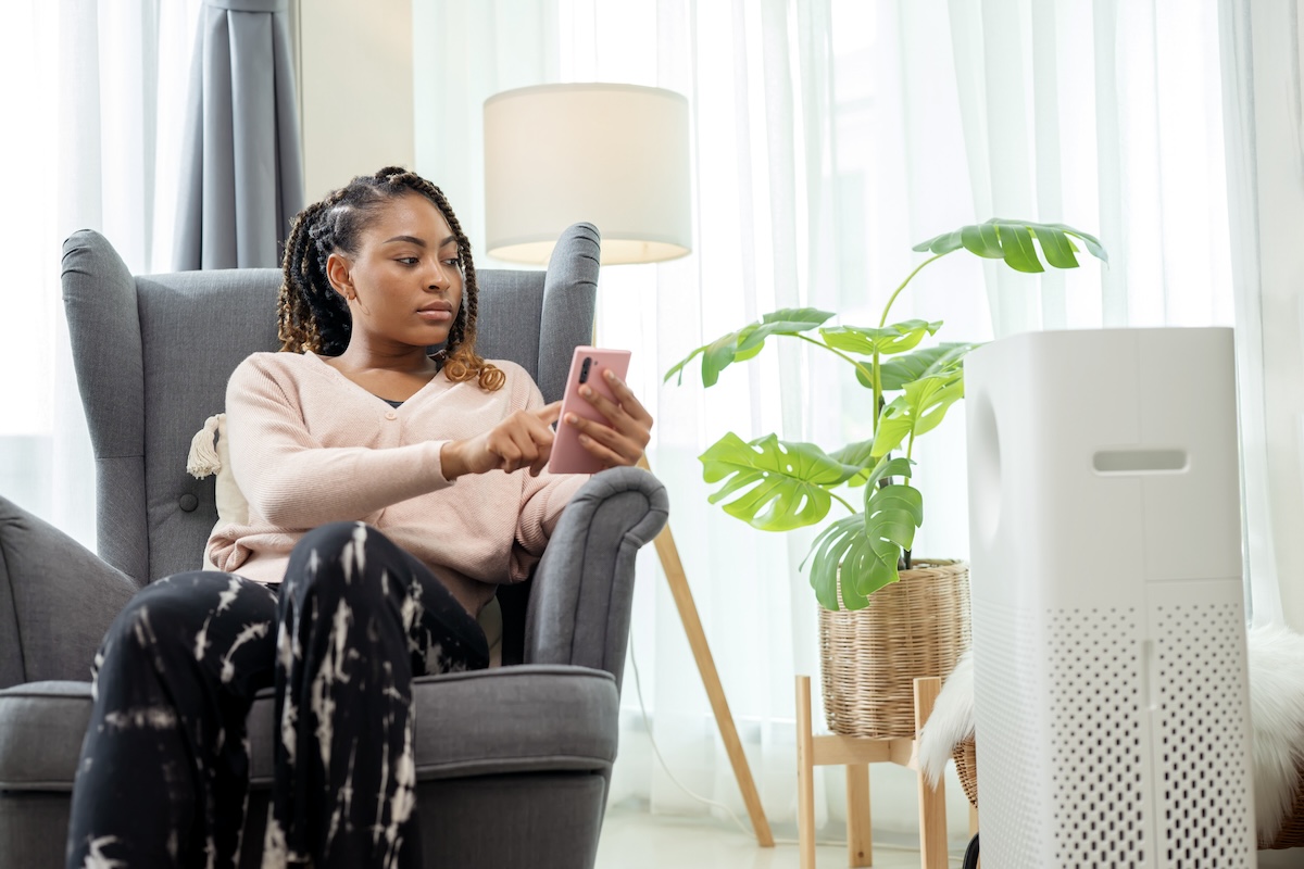 Home Automation Creates Comforting Routines and Increases Safety. An African American woman using a mobile app to turn on or off, adjust of an air purity machine in a living room at home.