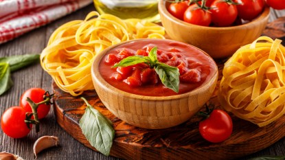 Bowl of tomato sauce on a wooden tray surrounded by pasta and fresh tomatoes