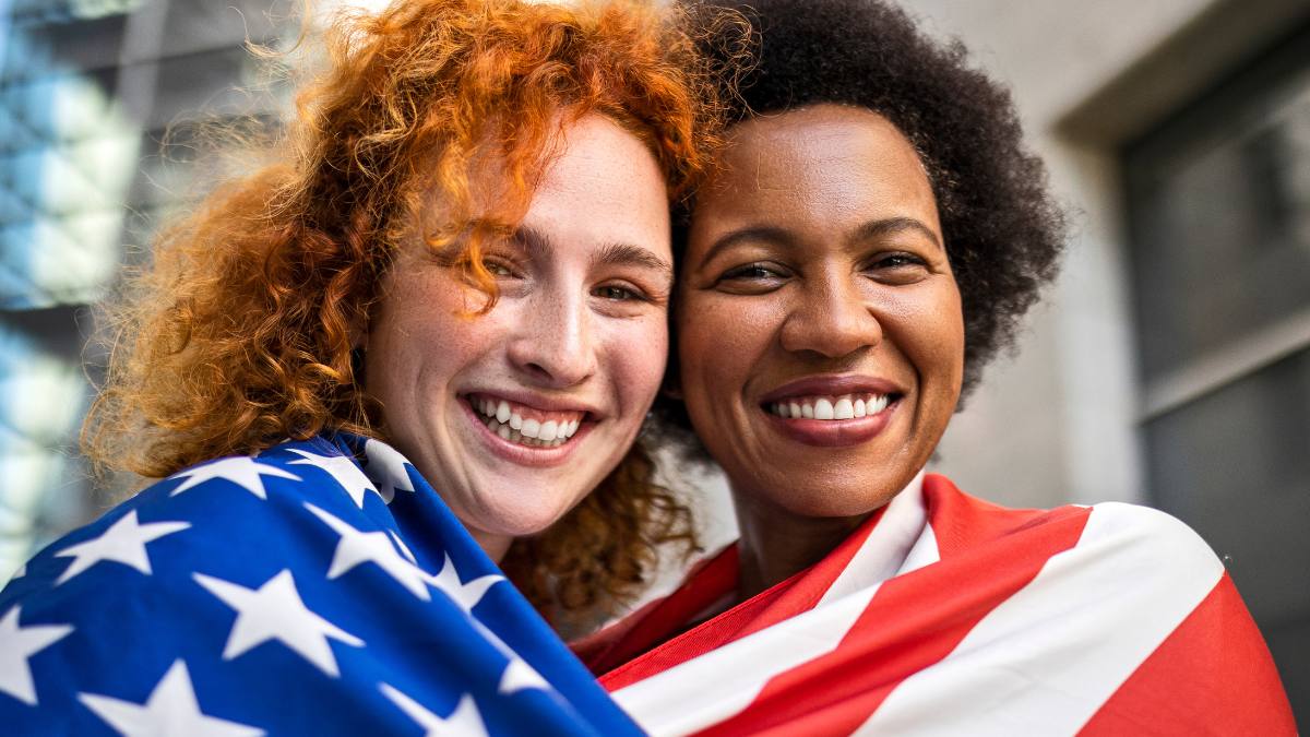 Girlfriends celebrating the 4th of July covered with American flag looking at camera