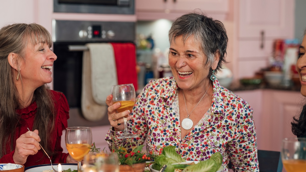 A shot of a small group of female friends gathered together in the kitchen of a home in Hexham, Northumberland. They are laughing joyfully whilst enjoying dinner together. The table is laden with bowls of colourful home-prepared food and wineglasses.