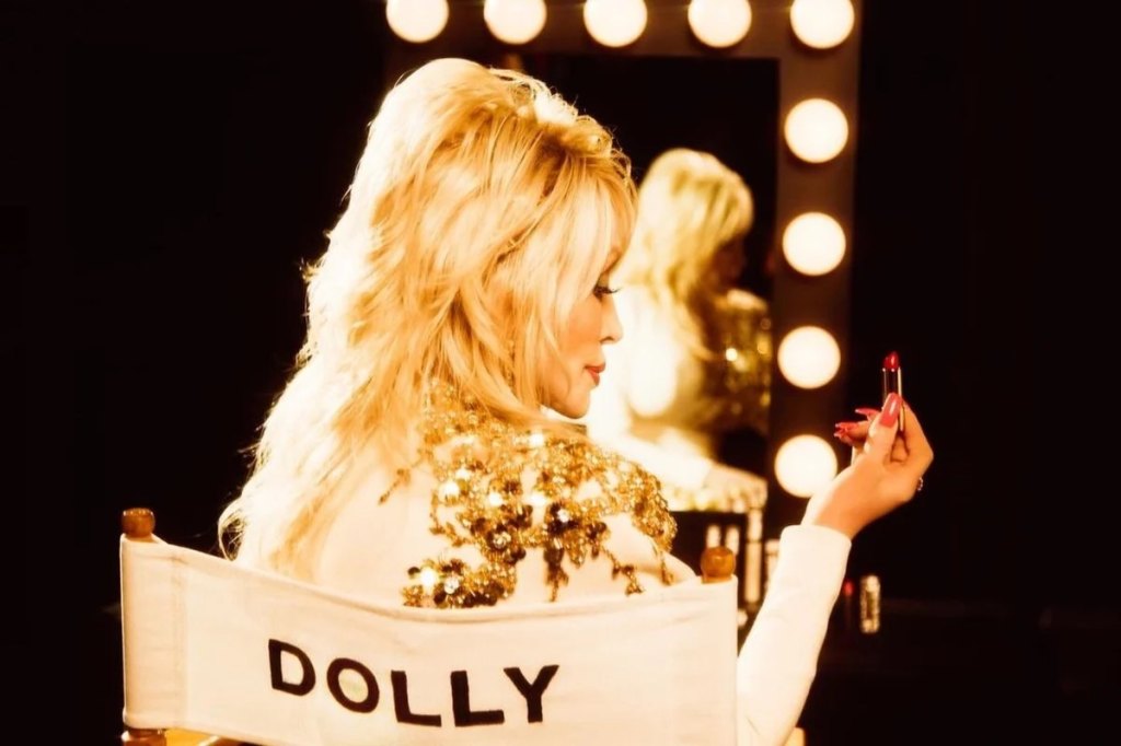 Dolly Parton sitting in a chair holding one of the Dolly Parton lipsticks