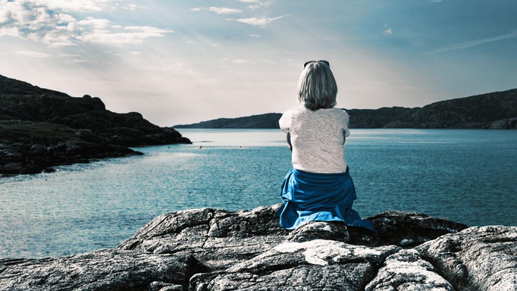 middle-aged woman, her back to us, sits on a rocky shore looking out at the horizon