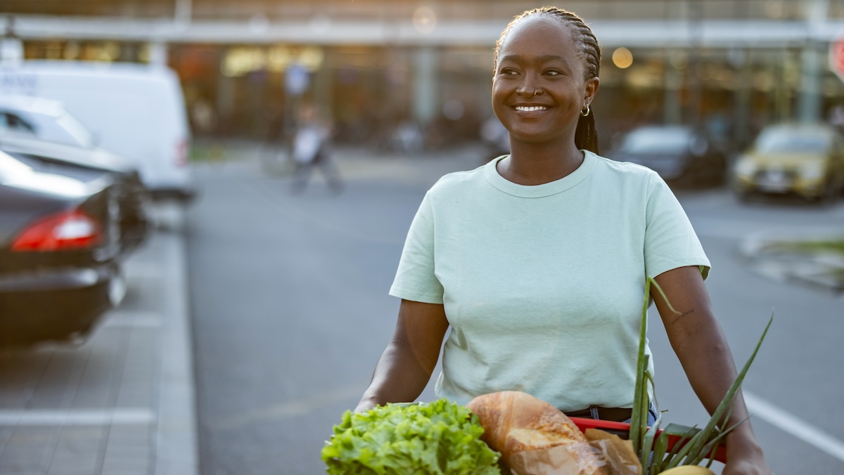 A cheerful woman with a cart full of groceries stands in a parking lot, her satisfied smile suggesting a successful shopping trip at the local supermarket.