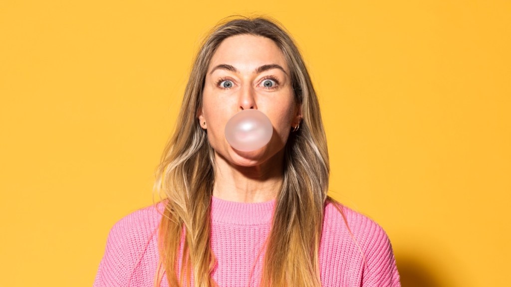 A woman in a pink shirt with dry mouth blowing a bubble with gum
