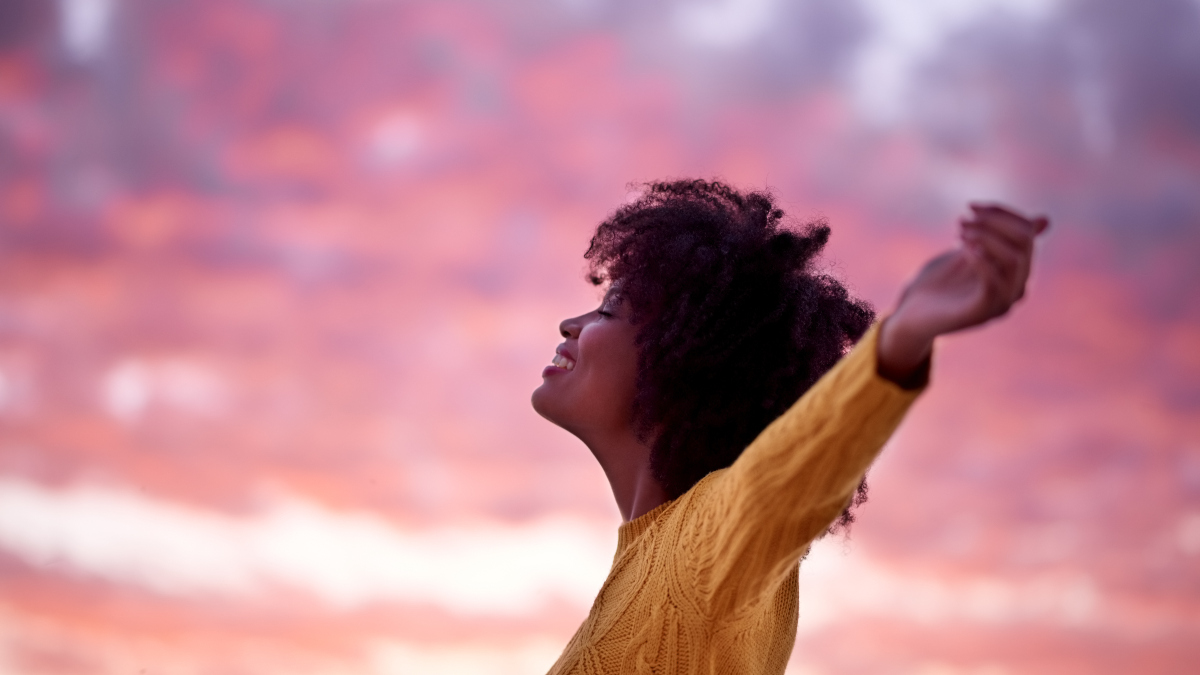 woman stretches out her arms, embracing a pink and purple sunrise, as she feels hopeful for the day ahead