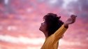 woman stretches out her arms, embracing a pink and purple sunrise, as she feels hopeful for the day ahead