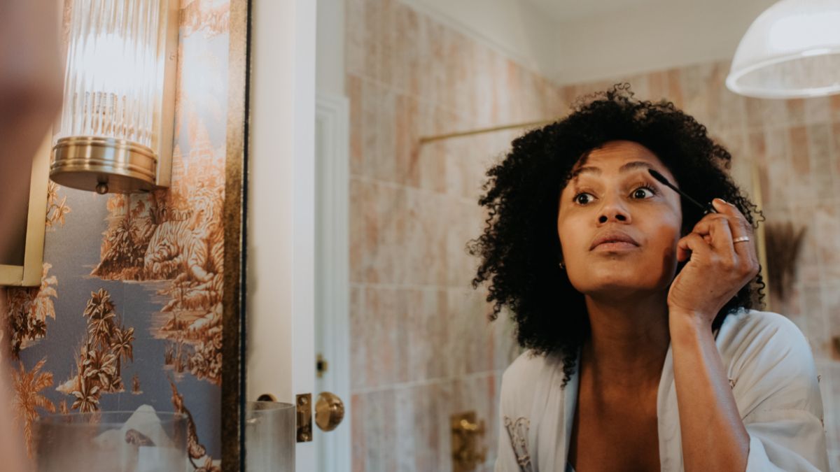 A beautiful young black woman concentrates as she applies mascara in a bathroom mirror.