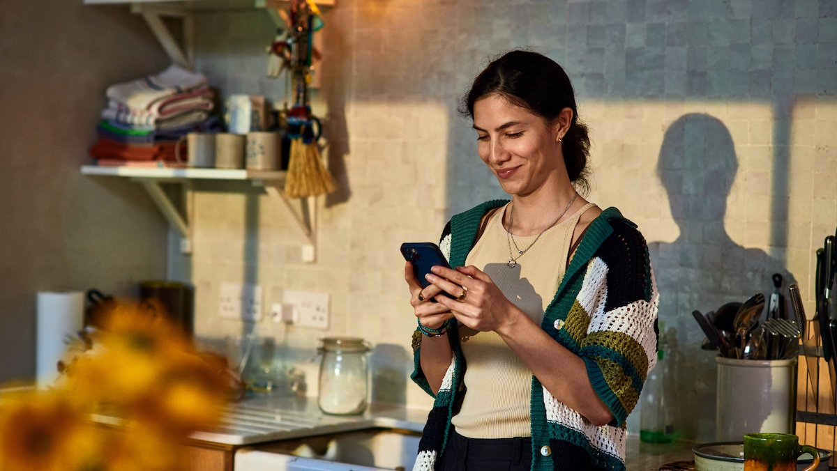 Smiling woman in her 30's standing in kitchen texting on smart phone, communication, wireless technology, the internet
