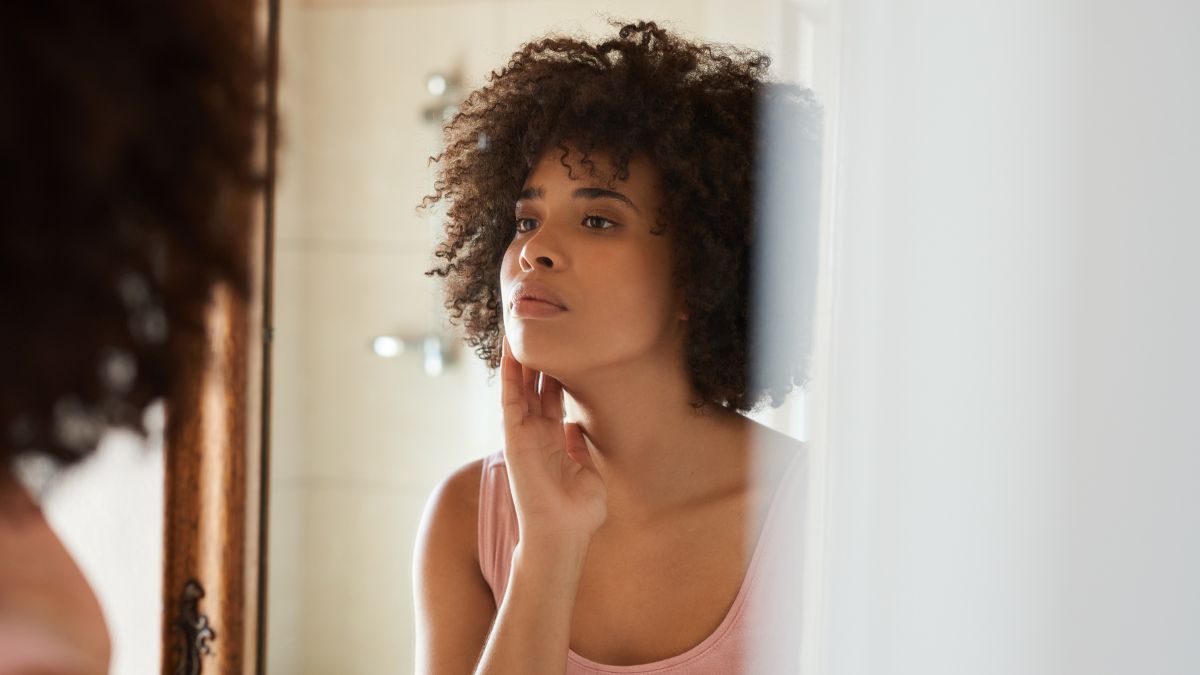 Young African woman examining her complexion in a mirror while standing in a bathroom at home in the morning