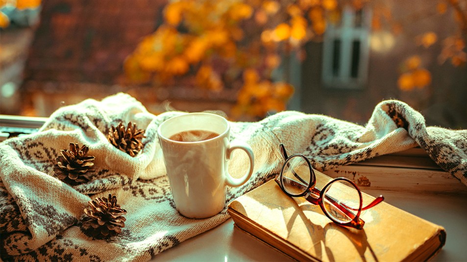 Cozy Fall Reads Feature Image: Mug, reading glasses and hot mug of tea on windowsill overlooking autumn trees