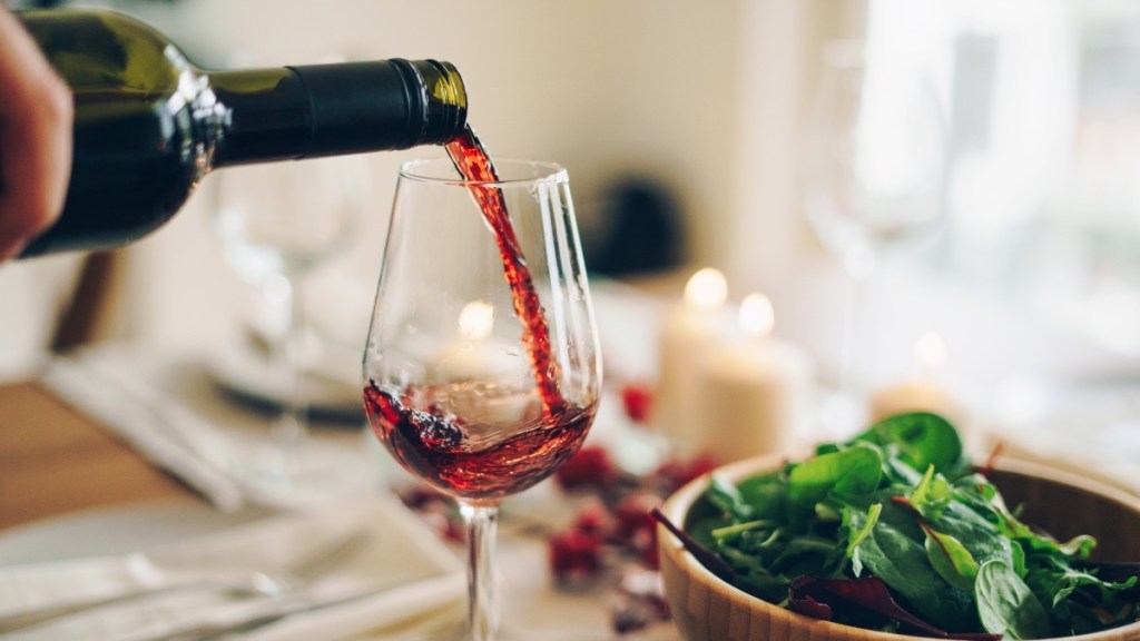 A bottle of red wine being poured into a clear glass beside a bowl of spinach