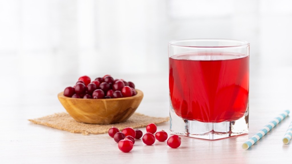 A glass of cranberry juice next to a bowl of cranberries and a blue straw