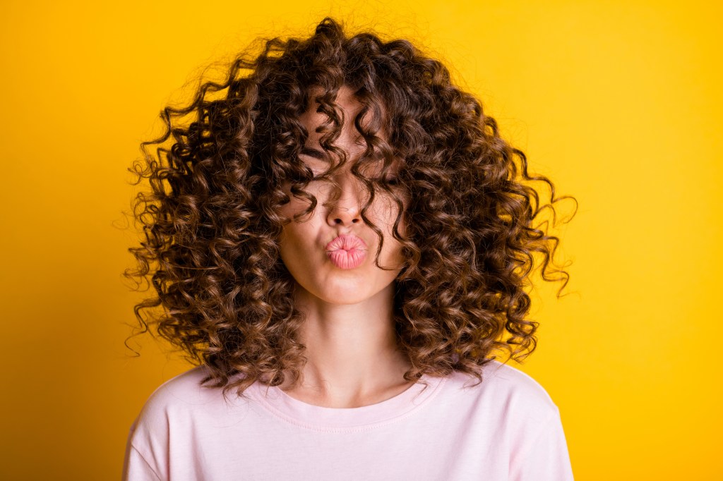 Woman with brunette curly hair in front of a yellow backdrop who is making a kissy face