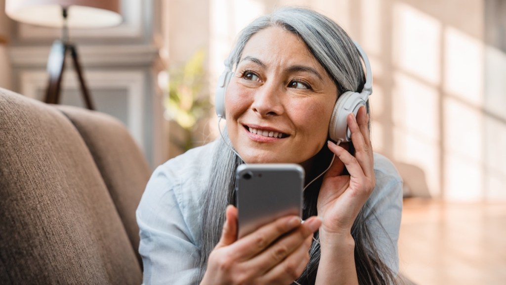 A woman with grey hair wearing headphones holding her phone listening to an audiobook