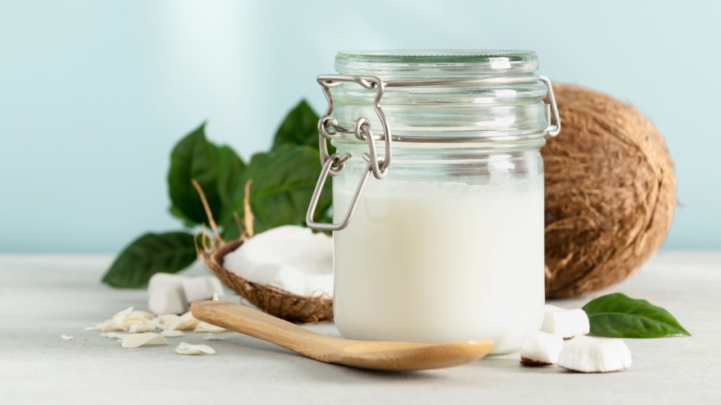 A jar of coconut oil next to a wooden spoon and cracked open coconut