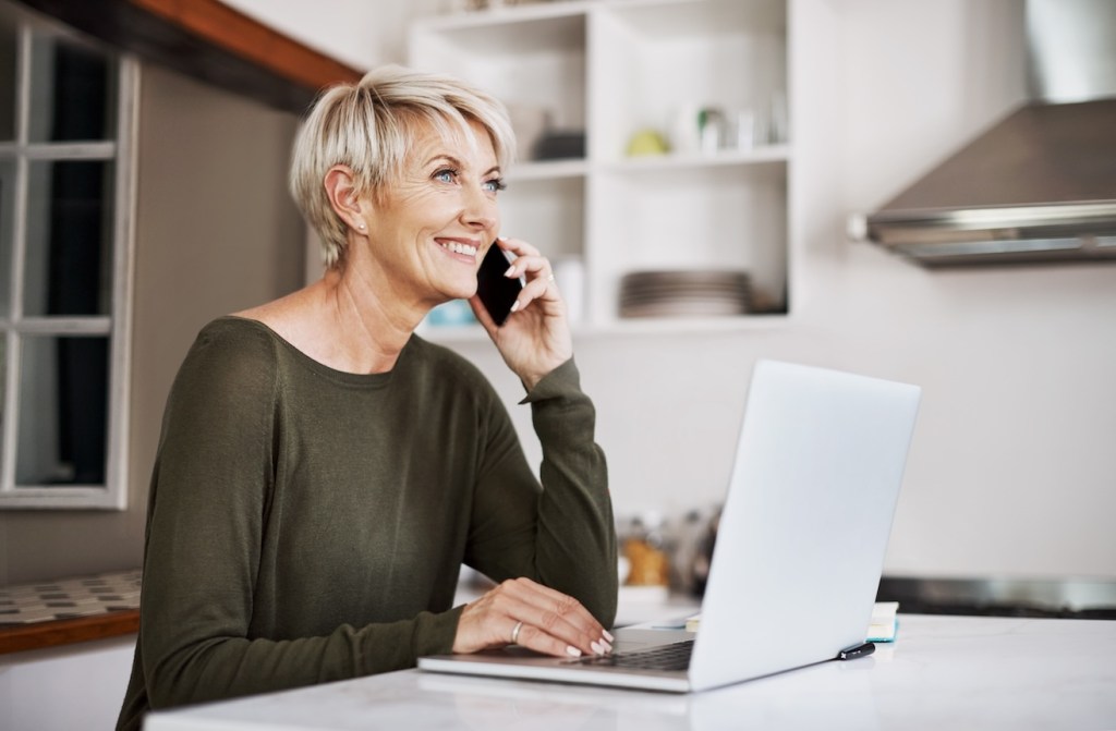 (CVS work from home) Shot of a mature woman talking on a cellphone while using a laptop at home