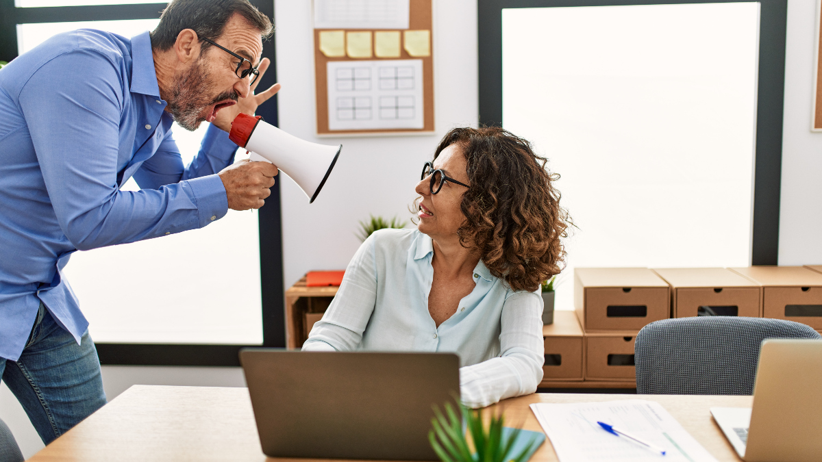 woman sits at her desk as her boss stands above her, yelling at her using a megaphone