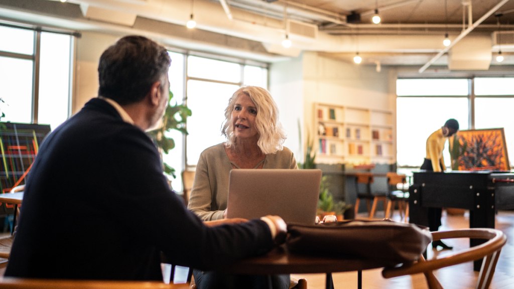 older woman with laptop open sits and talks with coworker