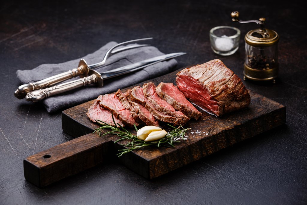 sliced roasted beef tenderloin on cutting board with knife and fork, salt and pepper and garlic